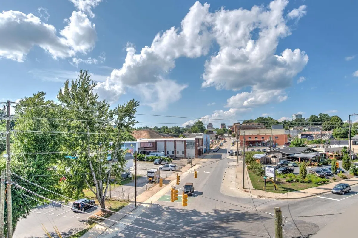 A lively street scene capturing the essence of Asheville’s urban energy. The image features commercial buildings, greenery, and traffic signals, with people walking and engaging in activities like dining and shopping. The overall mood is energetic and vibrant, reflecting the dynamic atmosphere of the neighborhood.