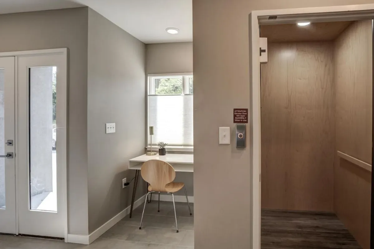 An interior shot featuring a desk with chair positioned near the elevator entrance and a glass door, highlighting a contemporary and functional entryway area. The space is clean, modern, and suggests ease of movement between different parts of the home.