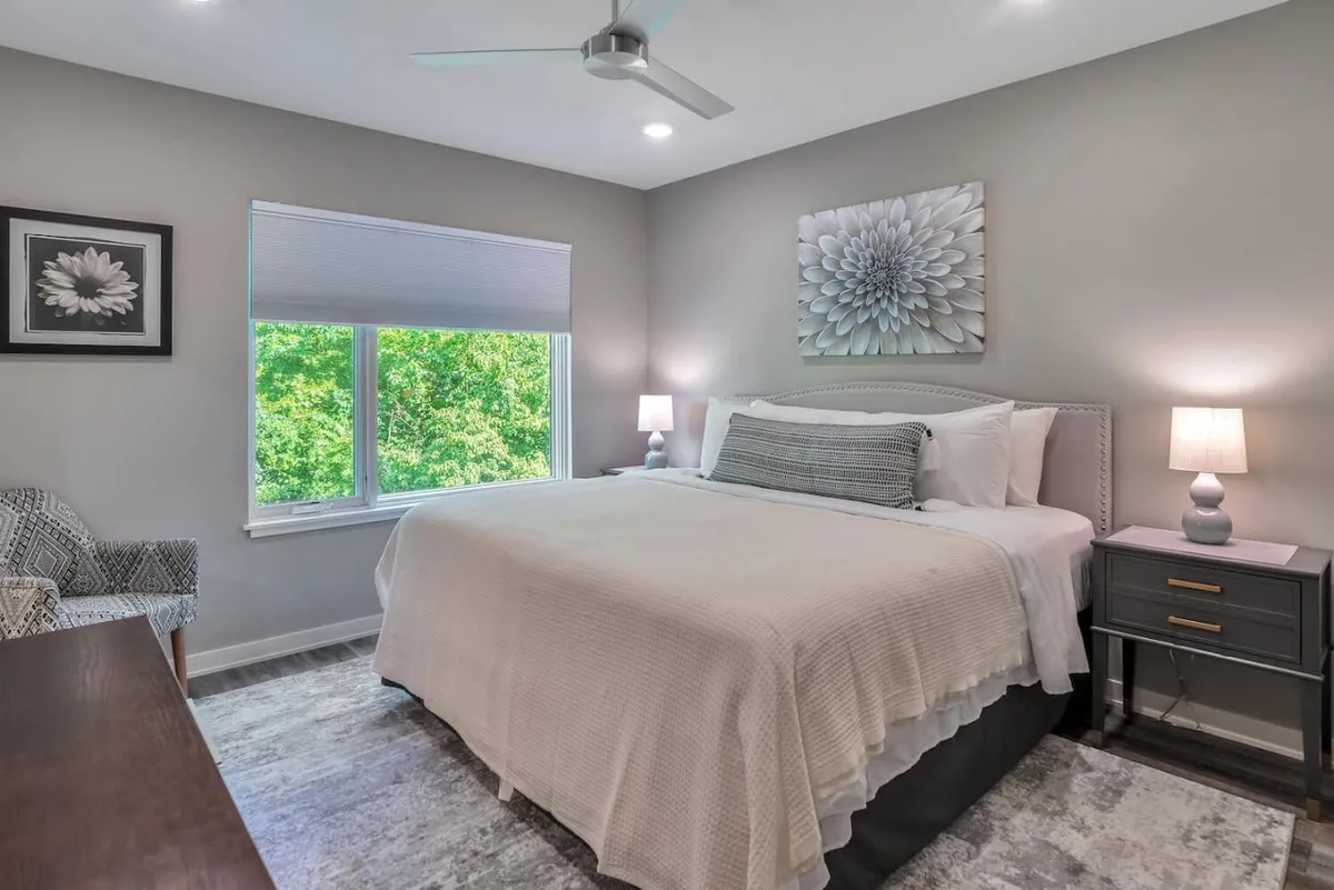 A serene king bedroom featuring a plush king-size bed with crisp linens, decorative wall art above the headboard, nightstands with lamps on either side, and a large window overlooking lush greenery. The soft natural light and cozy atmosphere make this space inviting for rest and relaxation.