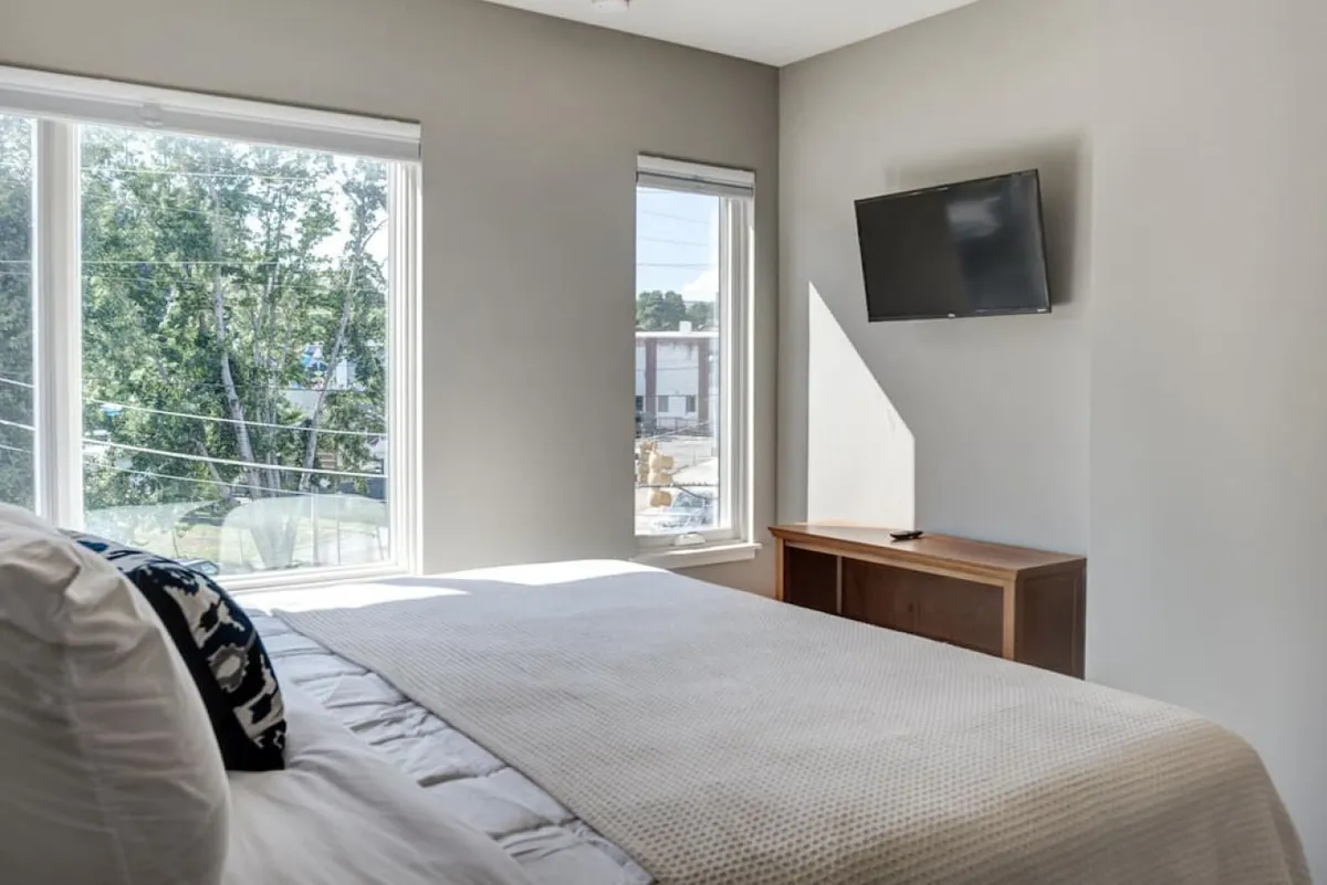 A serene bedroom with a neatly made bed featuring textured bedding, large windows letting in plenty of natural light, and a wall-mounted TV. The setting is modern and tranquil, ideal for restful nights or catching up on work in privacy.