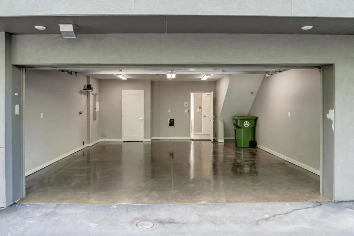 This image displays a clean, organized garage space with a green trash can and visible interior doors leading directly into the home. The area is practical and well-maintained, clearly serving as a secure parking and storage facility—ideal for arriving home after time spent in the city.