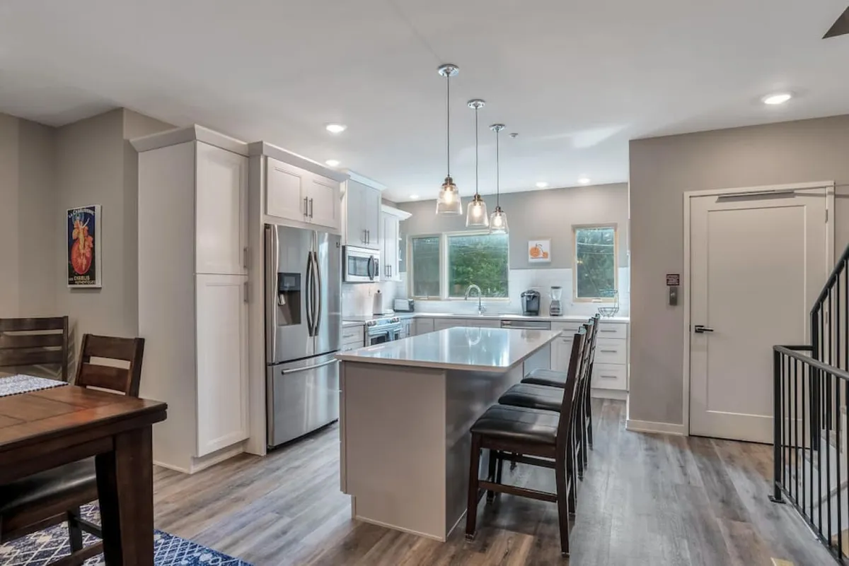 A bright, open kitchen area with an island countertop, modern appliances, pendant lights above the counter, and a dining table in the background. The overall layout is contemporary, emphasizing clean lines and a spacious, airy feel that invites group gathering and meal preparation.