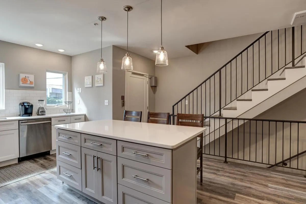 A modern, inviting kitchen scene with an island countertop, stainless steel appliances, and a staircase leading up from the background. The open-concept design allows for visibility between cooking and social areas, with ample space for entertaining guests or family.