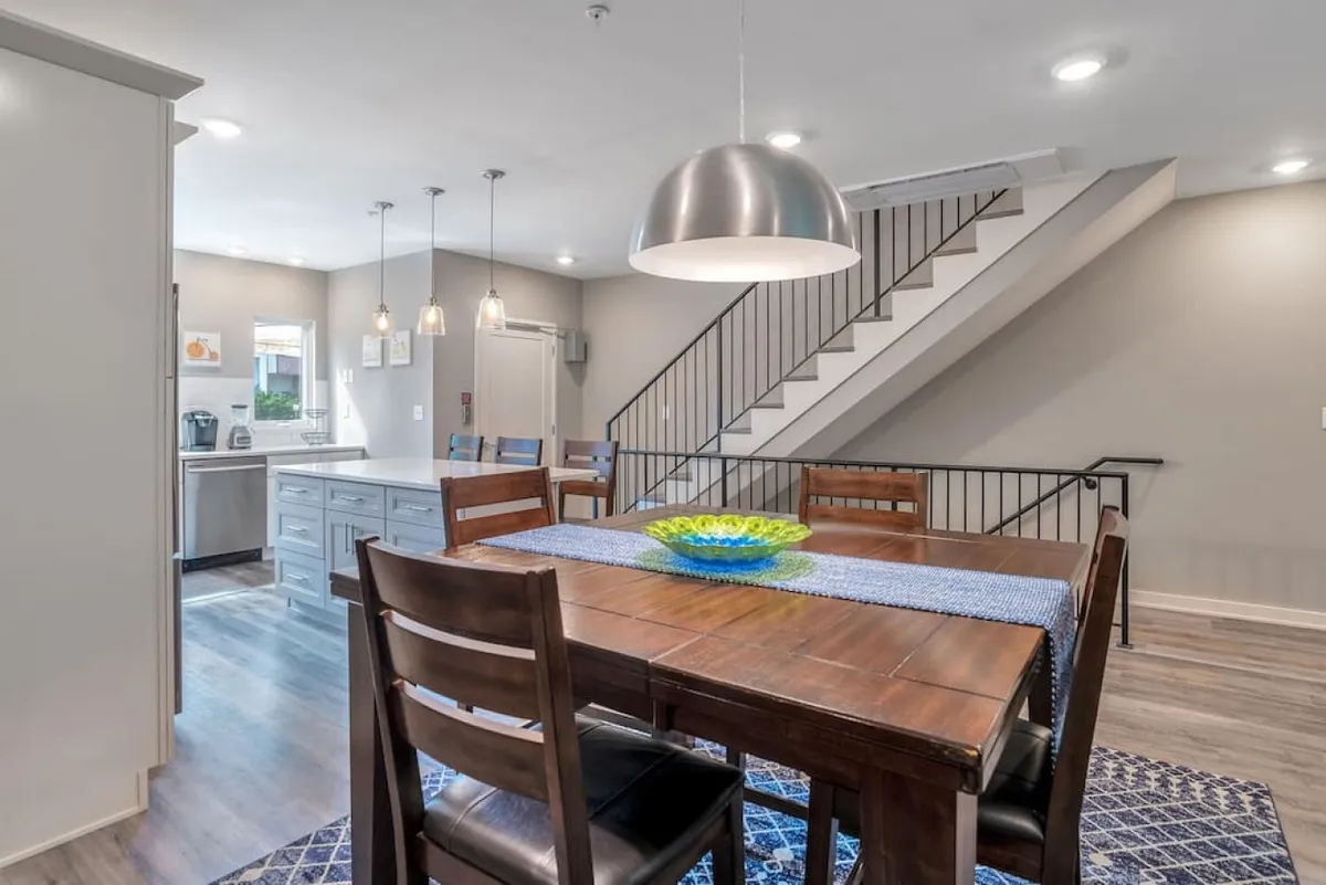 A well-lit dining area with a sizable table, a vibrant bowl centerpiece, and a staircase leading upwards. The space feels open and welcoming, ideal for shared meals or celebrations.