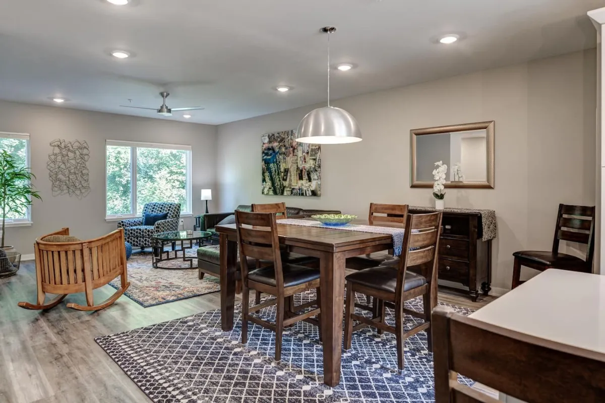 A welcoming living and dining space featuring a stylish dining table, a comfortable sofa, a rocking chair, and large windows allowing natural light to fill the room. Artwork on the wall and soft textures enhance the feeling of warmth and togetherness.
